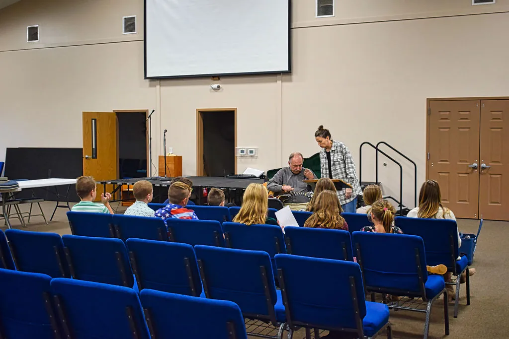 Children in rows of blue chairs listening to their teachers in the fellowship hall