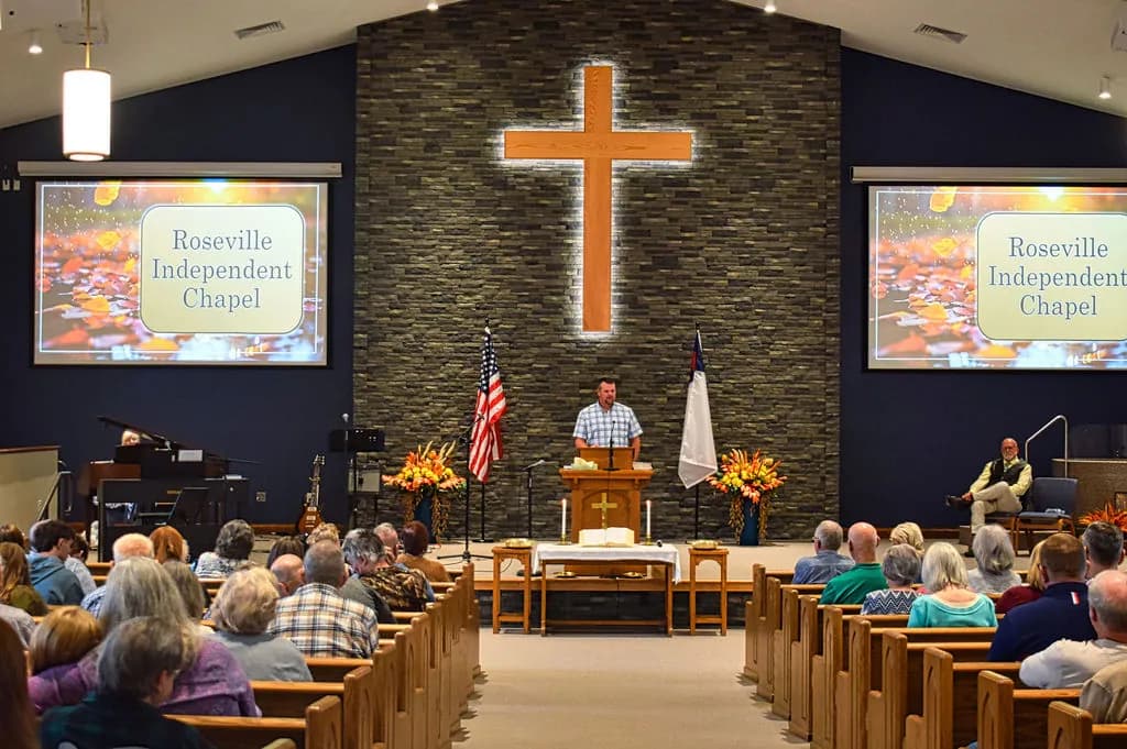 Sunday morning preaching at Roseville Independent Chapel, the lit wooden cross on the stone wall behind the pulpit
